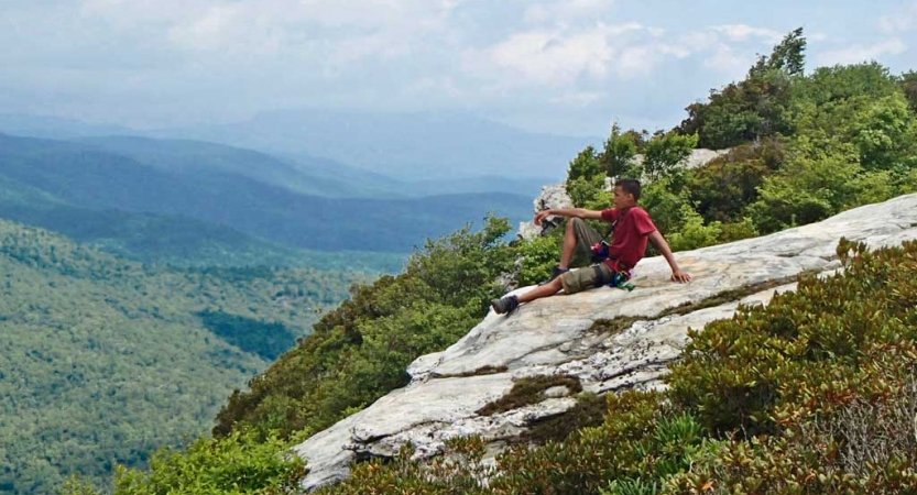 A student rests on a rock, overlooking a vast and green mountainous area. 